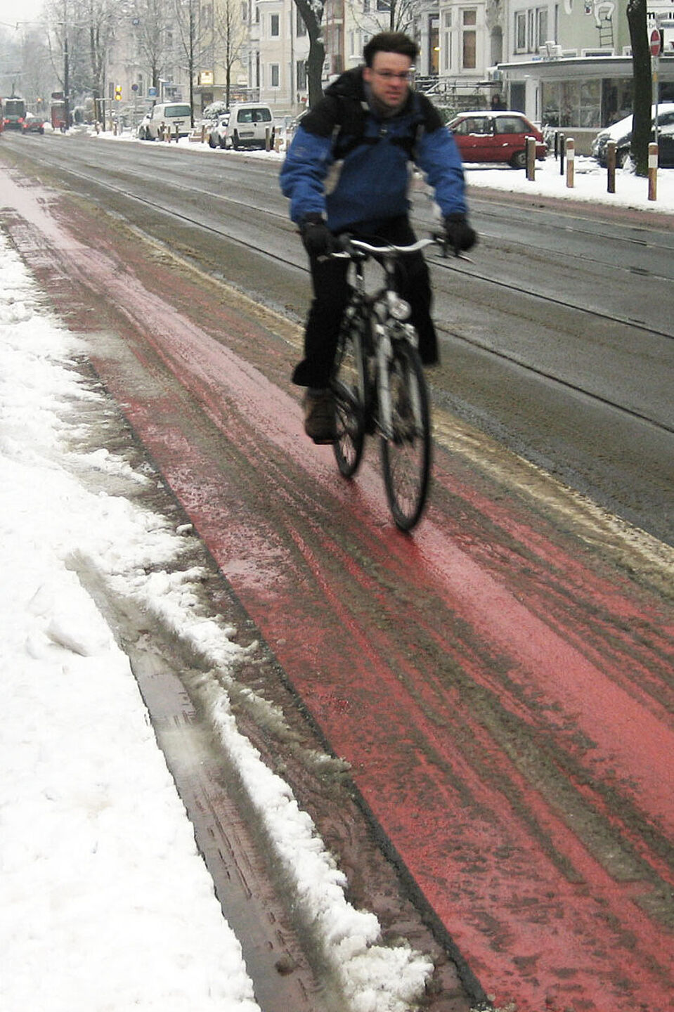 Radfahren im Winter Ein Radfahrer in Winterkleidung fährt auf einem rot markierten Radfahrstreifen neben einer Straße, der weitgehend von Schnee befreit ist, während am Straßenrand und auf geparkten Autos Schnee liegt.